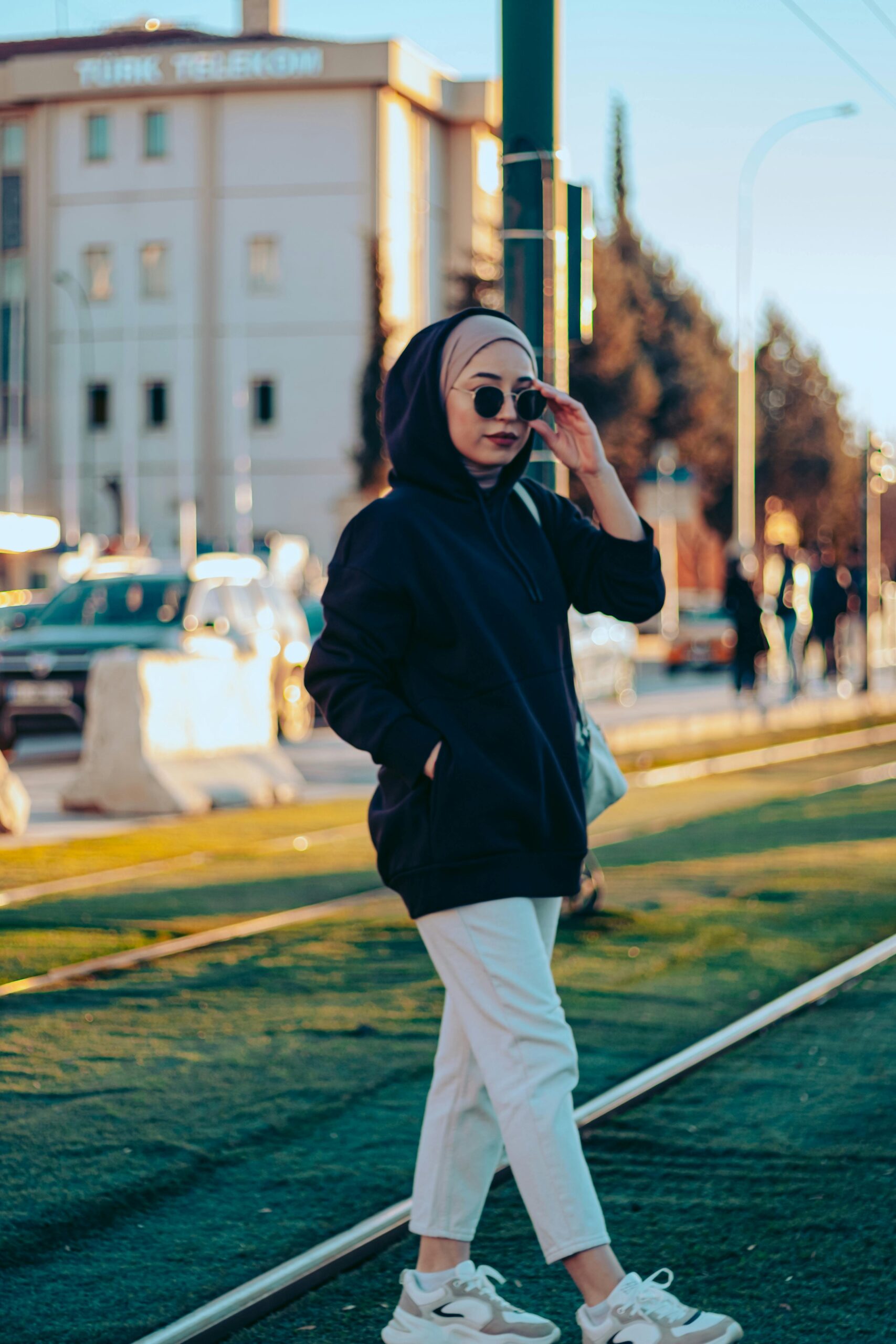 Woman wearing hijab and sunglasses, standing on a vibrant city street.