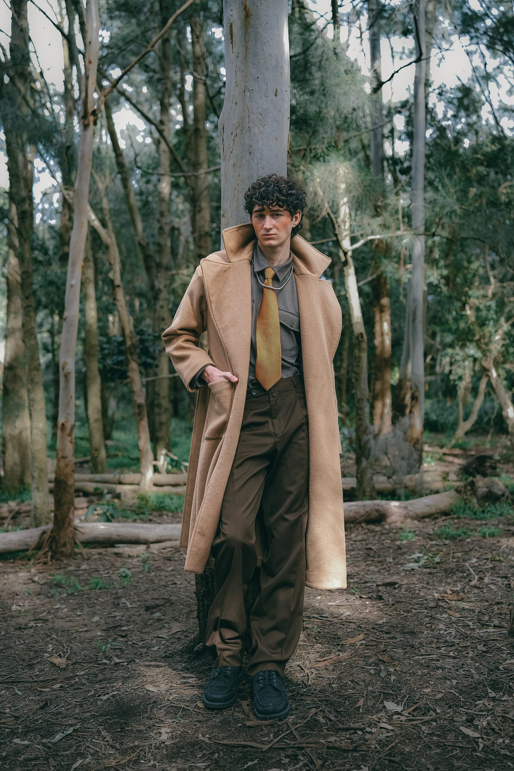 Man wearing a long coat and tie stands confidently in an autumn forest setting.